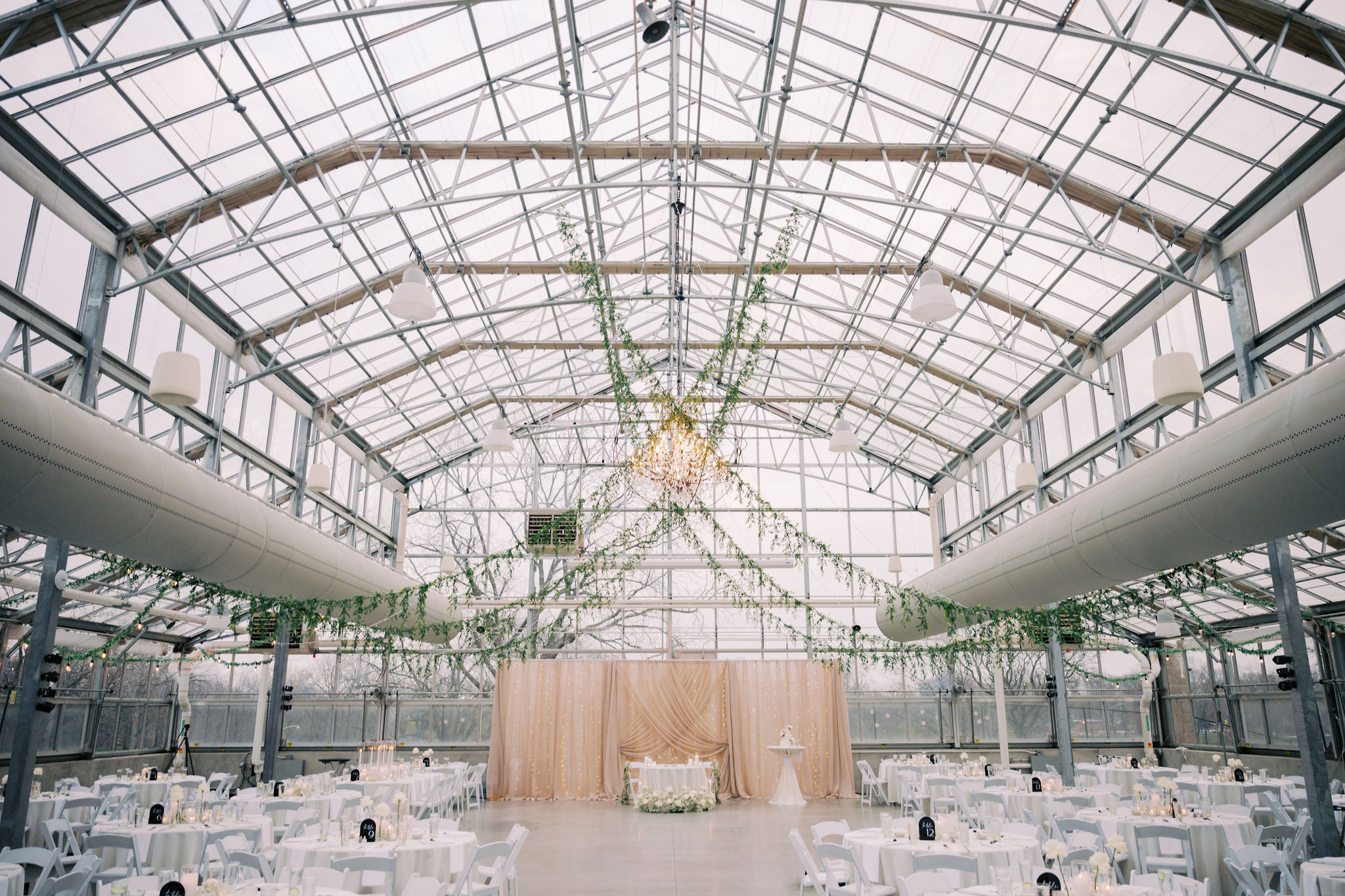 Large room with ceiling of windows and hanging greenery. Pink curtain backdrop and tables with white linens.